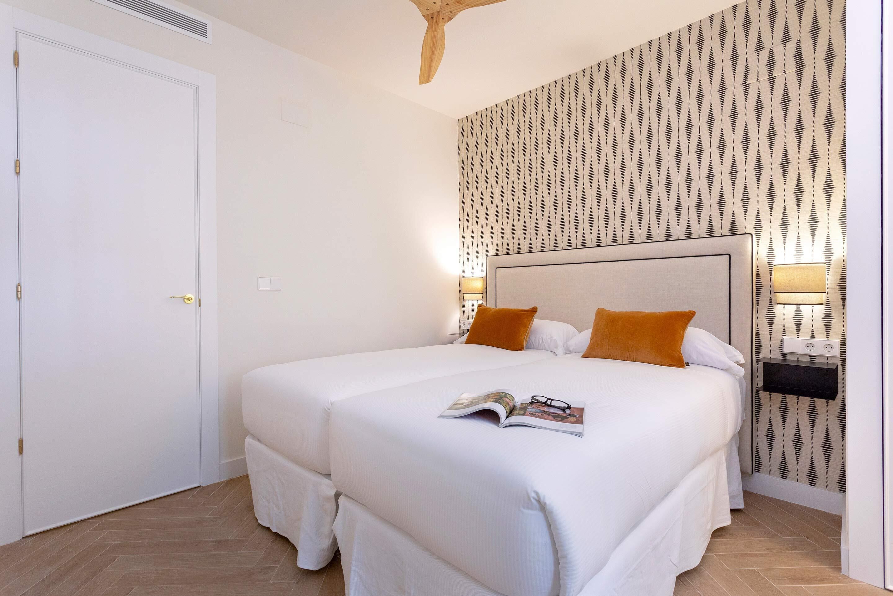 A double bed with white bedding and orange pillows, a bedside table with a lamp, and a patterned wallpaper on the wall.