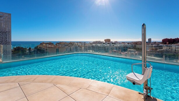 A clear blue swimming pool with a glass barrier, a sunshade, and a pool chair. - Genteel Home Infinity (Estepona)