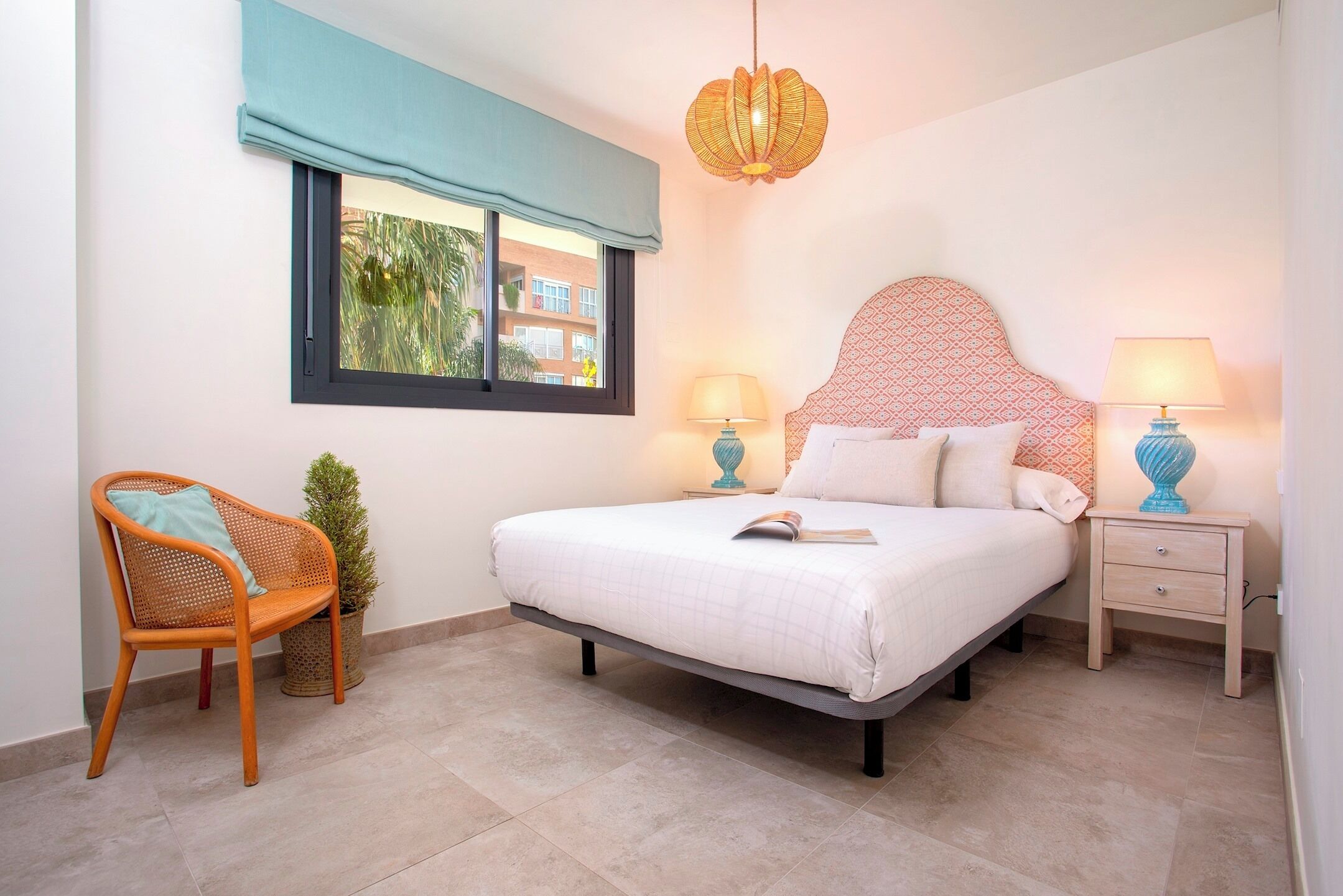 A bedroom with a bed, two bedside tables, a chair, a window with a view, and a hanging light fixture.