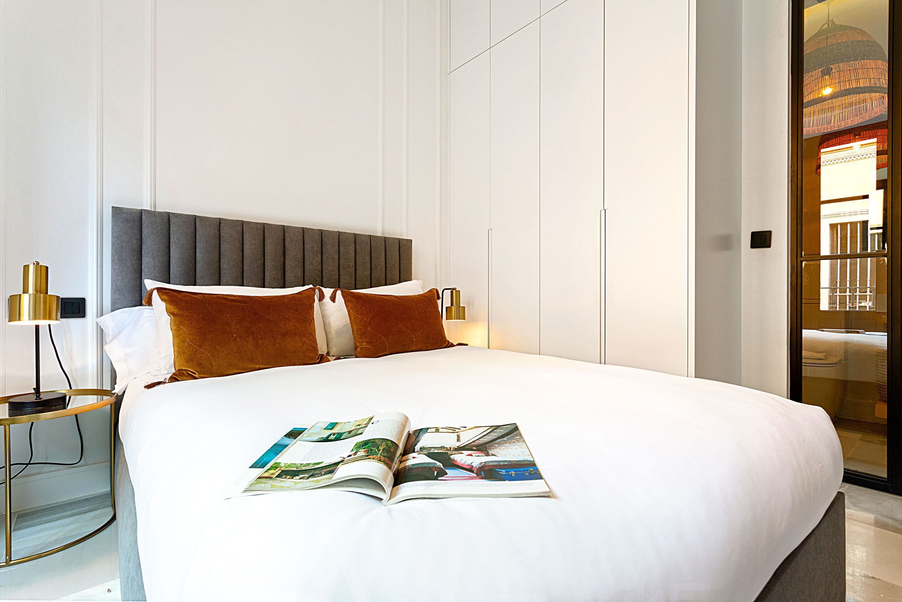 A neatly made bed with a magazine on it, a bedside lamp, and a view of a room with a large window and a hanging light fixture.