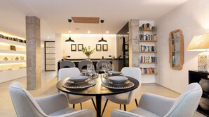 A modern dining area with a round table set for four, surrounded by light grey upholstered chairs. There are two pendant lights above the table, and a bookshelf filled with books is visible in the background. - Genteel Home Pureza (Seville)