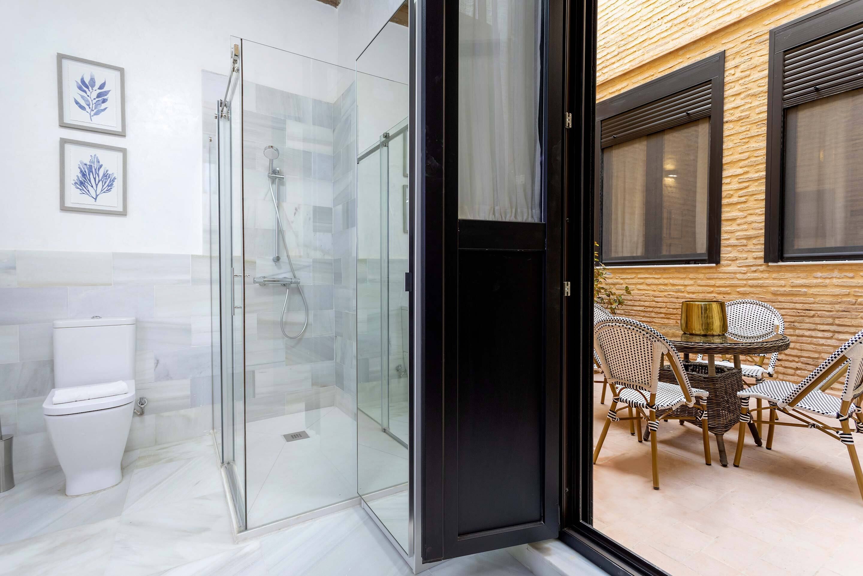 A modern bathroom with a glass shower enclosure, a toilet, and a dining area with wicker furniture and a table.