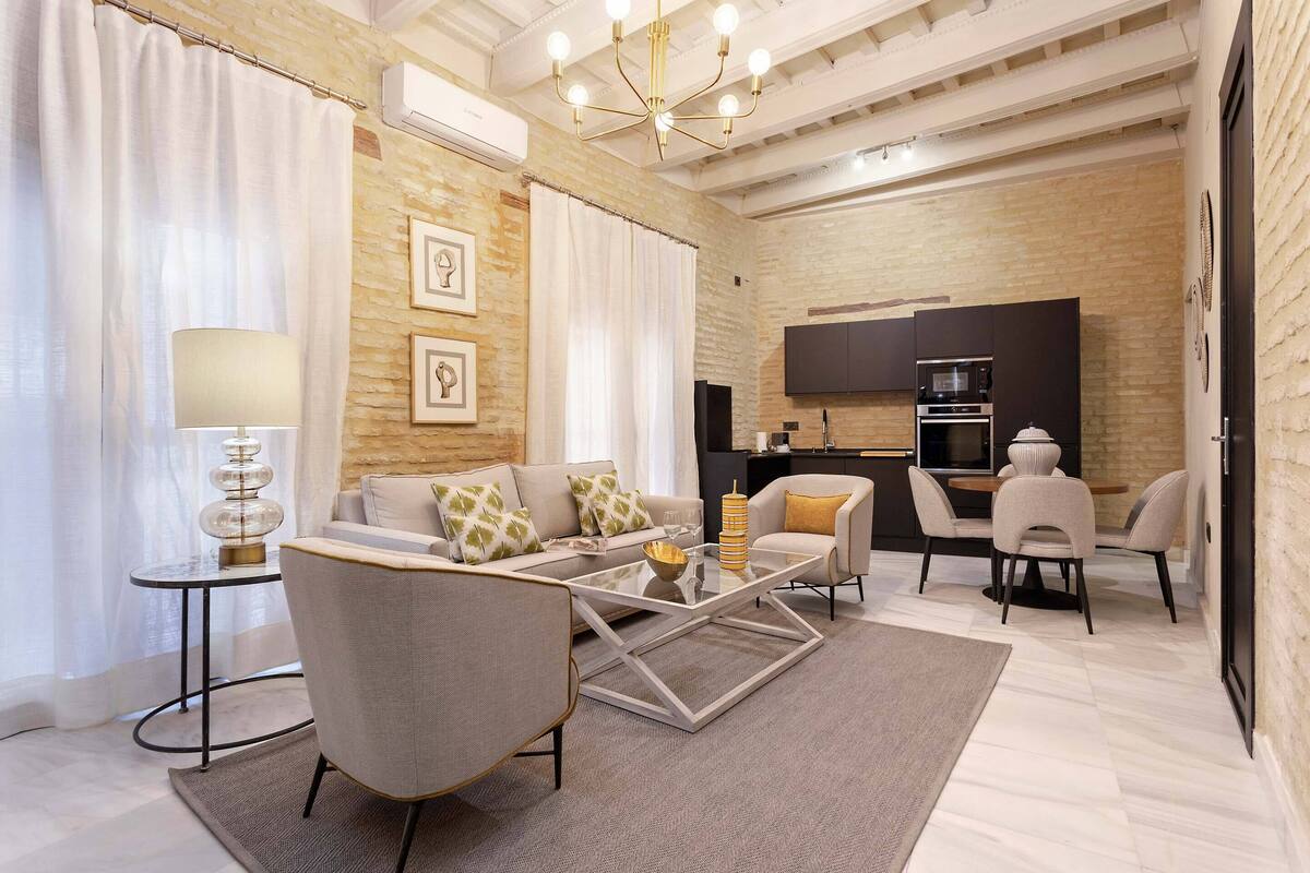 A modern living room with a sofa, armchairs, a coffee table, and a dining area with chairs. There is a kitchen in the background with appliances and a TV mounted on the wall.