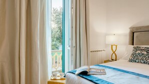 Una cama bien tendida con un detalle azul, una mesita de noche con lámpara y una ventana con cortinas.