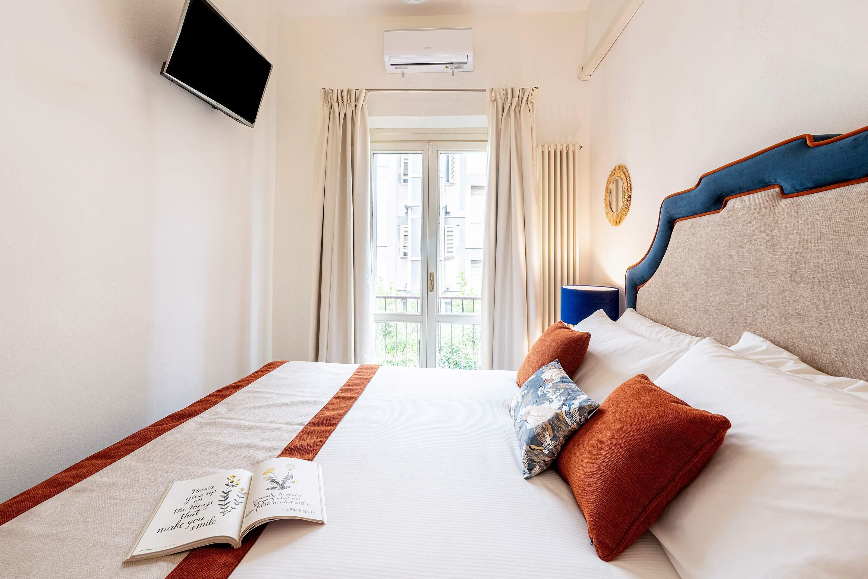 Une chambre d’hôtel avec un lit, un téléviseur à écran plat fixé au mur, une fenêtre avec des rideaux et une vue sur les bâtiments environnants.