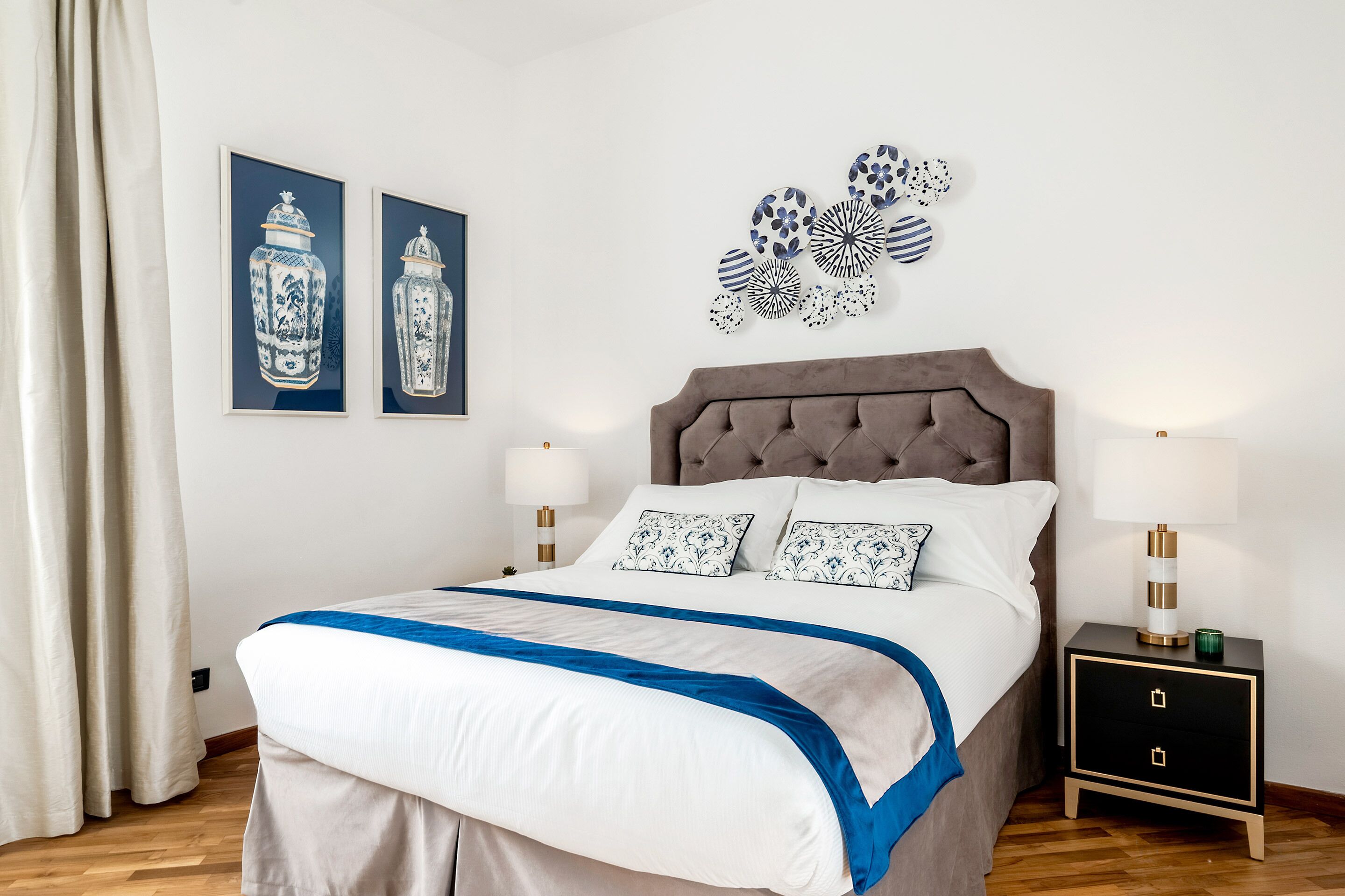 Una cama bien hecha con un detalle en azul, un cabecero capitonado y una mesita de noche con una lámpara.