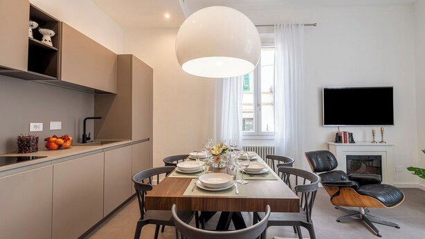 A modern dining area with a wooden table set for four, surrounded by chairs. A large pendant light hangs above the table, and a flat-screen TV is mounted on the wall to the right. - Genteel Home Cittadella (Florence)