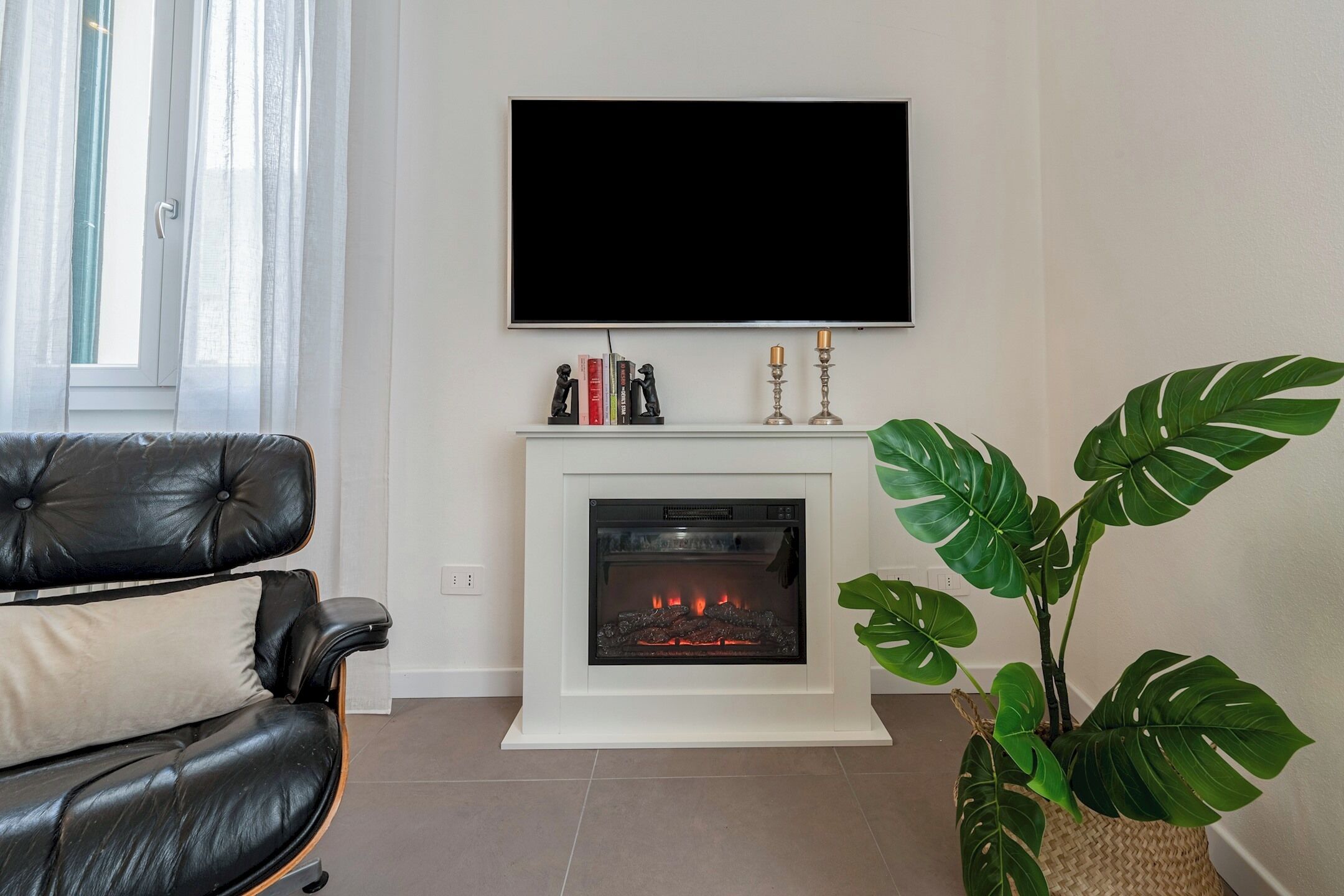 Una sala de estar con un sofá de cuero negro, una chimenea blanca, un televisor de pantalla plana montado sobre la chimenea y una planta verde grande.