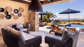 Un patio iluminado por el sol con mobiliario de mimbre, un pilar de piedra y vistas a una piscina y al océano.