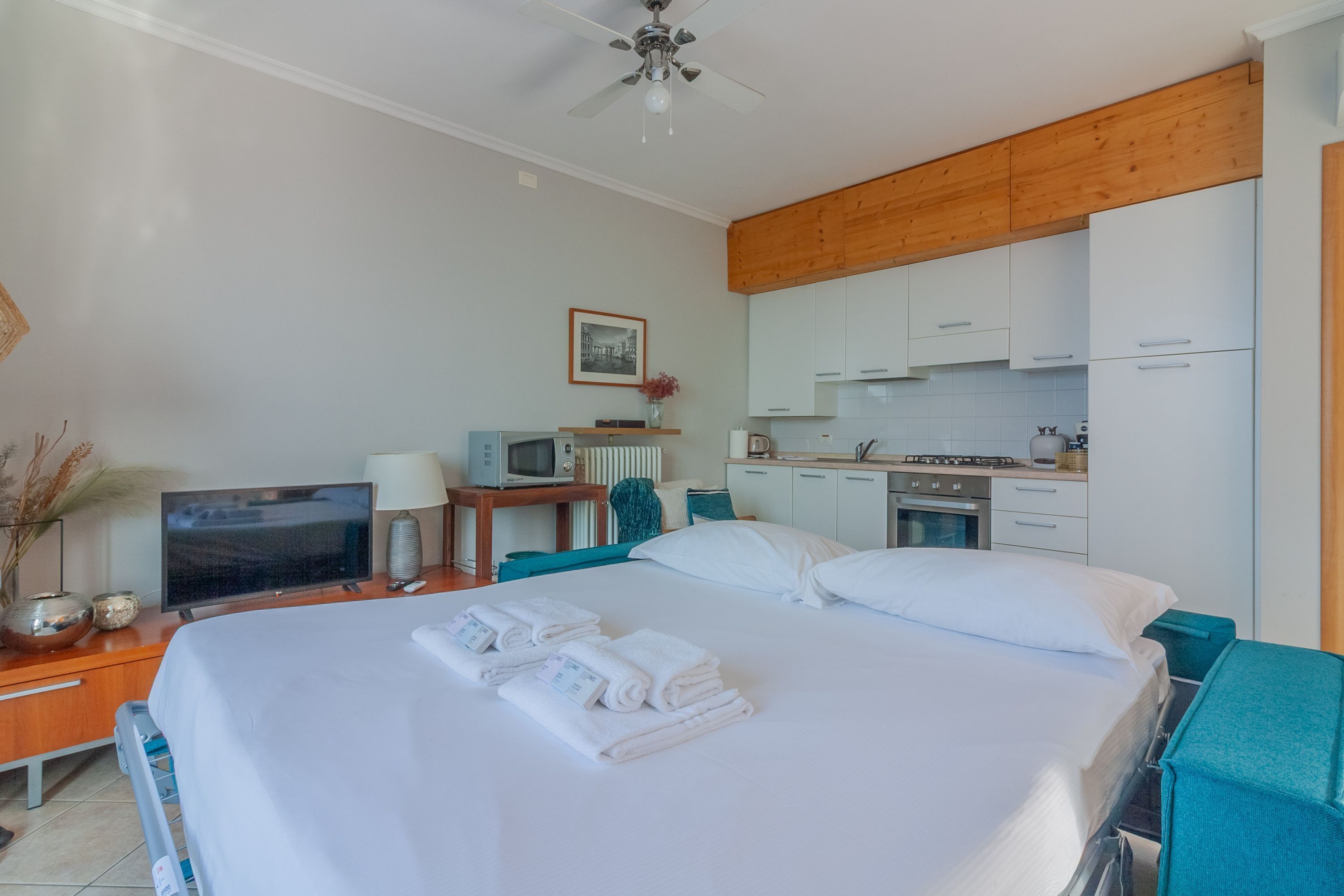A hotel room with a bed, a desk with a TV, a kitchenette, and a ceiling fan.