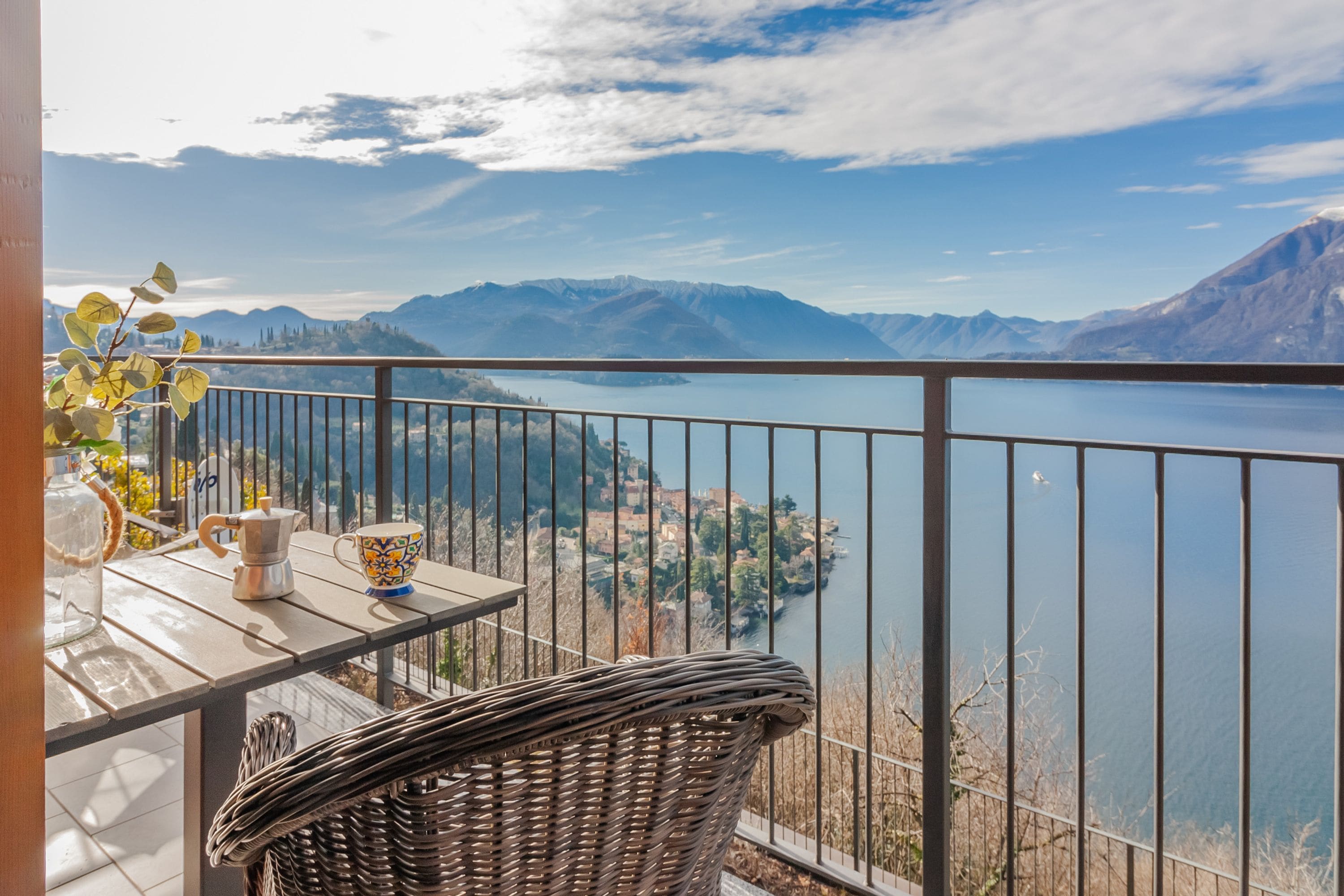 Un balcon donnant sur un lac et des montagnes, une chaise en osier, une table avec une tasse et un pichet, et une balustrade.