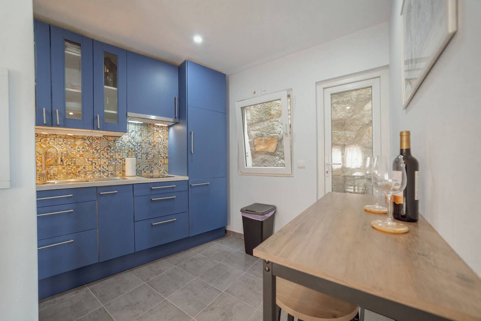 A kitchen with blue cabinets, a tiled backsplash, and a dining table with wine glasses and a bottle.