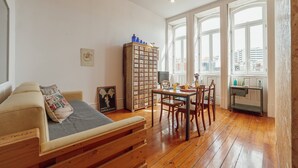 A cozy living room with a wooden sofa, a dining table with chairs, a tall bookcase, and a view through large windows. - Downtown Bolhão 3 by Hosty (Porto)