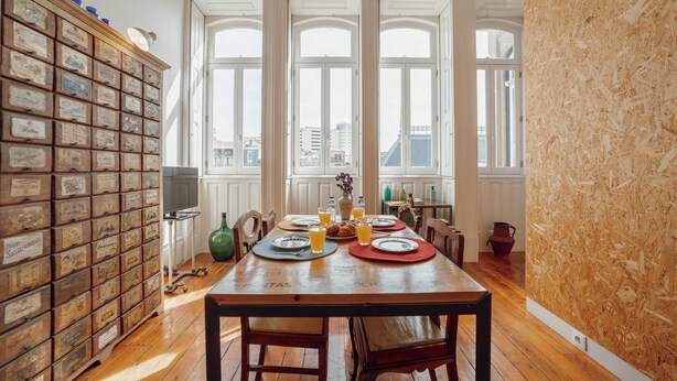 A dining area with a wooden table set for four, large windows, and a collection of vintage metal containers on the left. - Downtown Bolhão 3 by Hosty (Porto)