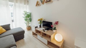 Un salon moderne avec un canapé gris, un meuble TV en bois sur lequel est posée une télévision à écran plat, une plante en pot et une petite table avec une lampe.