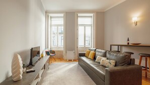 Una sala de estar moderna con un sofá, una mesa de centro y un televisor. Hay dos ventanas con persianas, un radiador y un accesorio de iluminación fijado a la pared.