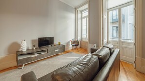 Una sala de estar moderna con un sofá gris, un televisor sobre un mueble bajo de madera, y una ventana con vistas a edificios.