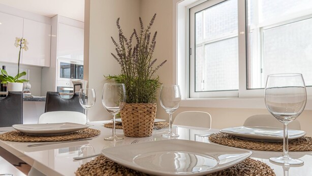 Een moderne eetruimte met een lange tafel gedekt voor vier personen, voorzien van geweven placemats, witte borden en wijnglazen. Een potplant zorgt voor een vleugje groen.