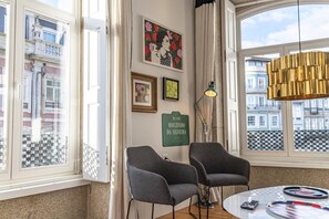Une pièce avec deux fauteuils gris, une table ronde avec une télécommande et un jeu de cartes, un panneau vert et une grande fenêtre donnant sur des immeubles.
