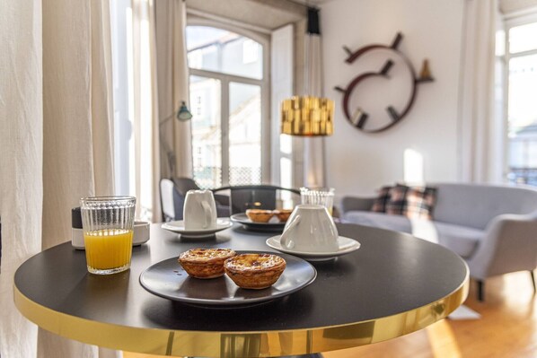 Una mesa de desayuno con pasteles y un vaso de jugo de naranja, en una habitación luminosa con un ventanal grande y un reloj en la pared.