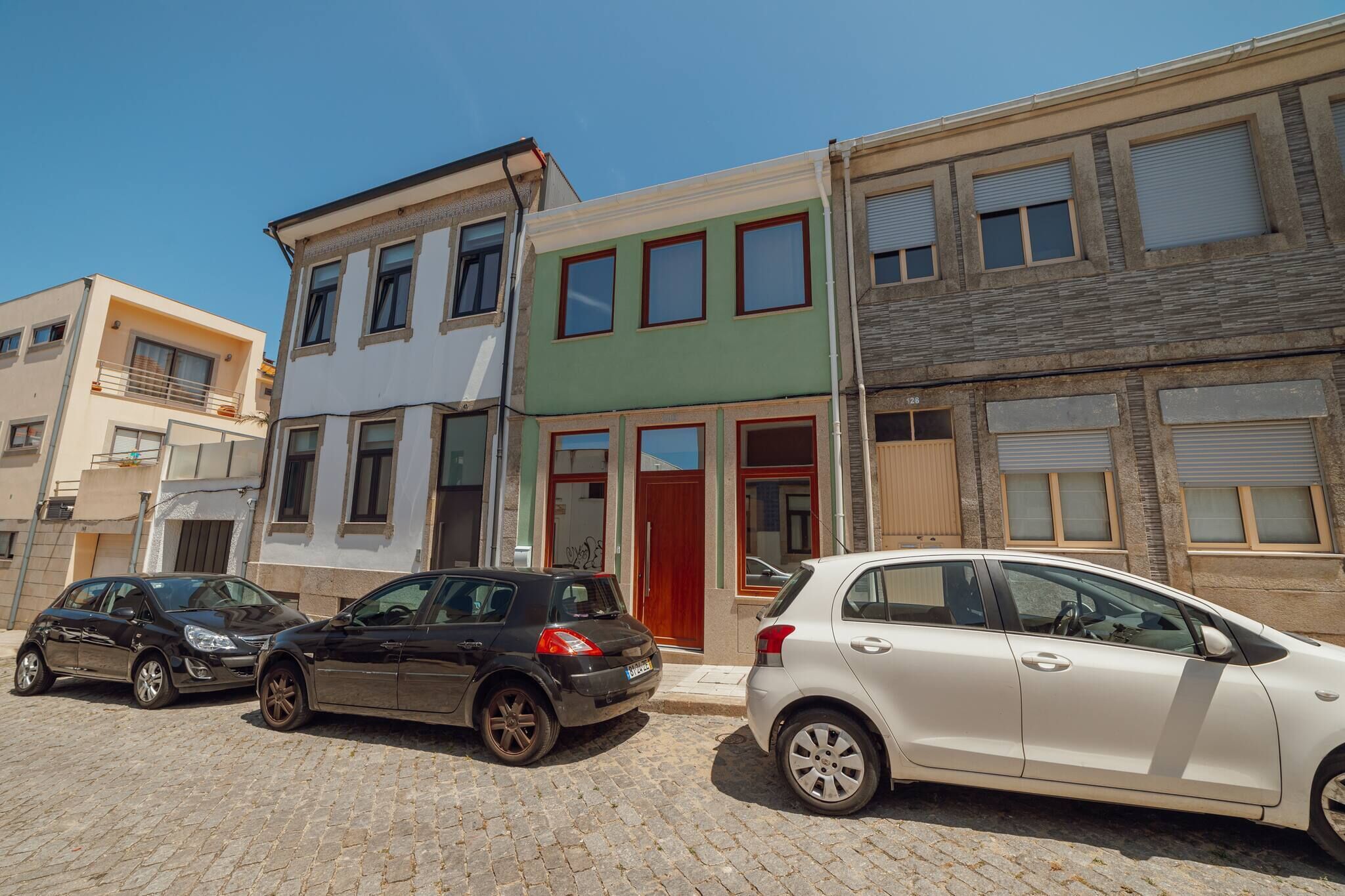 Uma fileira de edifícios de três andares com uma porta vermelha e janelas. Dois carros estão estacionados em frente.