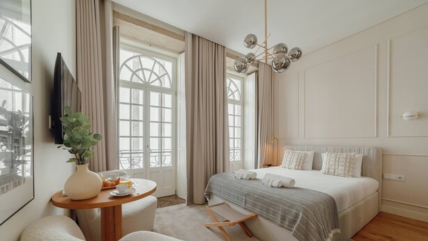 Un dormitorio moderno con una cama grande, una mesita de noche con un florero y fruta, y vistas de la ciudad a través de amplias ventanas.