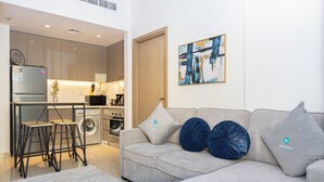 A modern living room with a grey sofa, two blue decorative pillows, a grey rug, and a kitchen area with appliances. - Bloomfields Modern 2BR Azizi Gardens (Dubai)