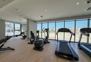 A gym with treadmills, elliptical machines, and stationary bikes, large windows with a city view. - Bloomfields 1BR+ 1 Azizi Gardens - Meydan (Dubai)