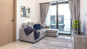 A modern living room with a grey sectional sofa, a patterned rug, a painting on the wall, and a balcony with a view. - Bloomfields Modern 2BR Azizi Gardens (Dubai)