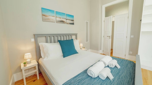 A neatly made bed with a blue pillow, a small bedside table with a lamp, and a rolled towel on the bed. - Bonfim Premium by Hosty (Porto)