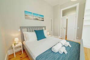 A neatly made bed with a blue pillow, a small bedside table with a lamp, and a rolled towel on the bed. - Bonfim Premium by Hosty (Porto)