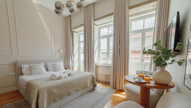 Ett modernt hotellrum med en säng, ett runt träbord med en vas och en kopp, och ett stort fönster med gardiner.