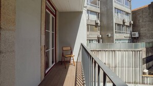 Un balcón con una silla de madera, un ventanal con marco rojo y vista a un edificio de varios pisos con unidades de aire acondicionado.