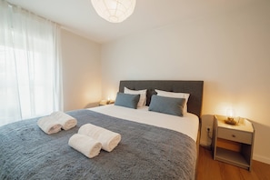 Een opgemaakt bed met kussens, een bedlampje en opgerolde handdoeken op een grijs geweven sprei.