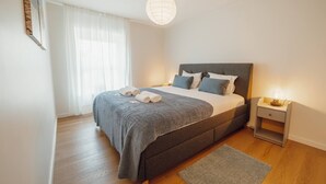 A bedroom with a bed, a nightstand, a lamp, a window with curtains, and a wooden floor. - Matosinhos Premium by Hosty (Senhora da Hora)
