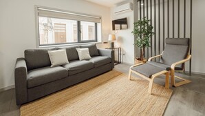 A modern living room with a grey sofa, a wooden chair, a small table with a lamp, a potted plant, and a window with blinds. - Amazing Villa by Hosty (Oporto)