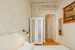 A bedroom with a white bed, a white wardrobe, a mirror, and a door leading to another room.