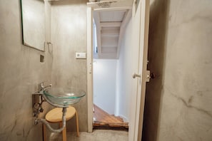 A small bathroom with a glass sink, wooden stool, and a narrow hallway leading to a staircase.