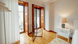 A room with a wooden floor, a chair, a small white table with a lamp, and a door leading to an outdoor area. - Bonfim Vintage by Hosty (Porto)