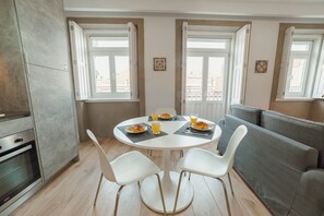 Un coin repas comprenant une table ronde, quatre chaises, un canapé et deux fenêtres avec des rideaux ouverts.