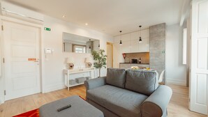 Een moderne woonkamer met een grijze bank, een klein voetenbankje, een salontafel, een plant en een keuken op de achtergrond.