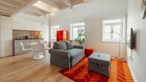 Een moderne woonkamer met een grijze bank, een kleine tafel en een rood vloerkleed. Er is een keuken met witte kasten en een eettafel met stoelen.