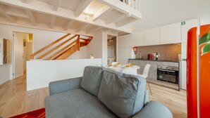 Een modern interieur met een grijze bank, een witte eettafel met oranje fruit, een keuken met een kookplaat en een trap.