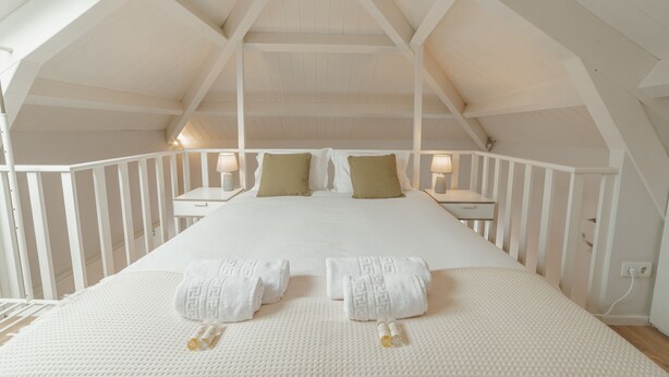 Een slaapkamer met een hoge plafond, witte muren en een houten plafondconstructie. Er staan twee nachtkastjes met lampen, een bed met wit beddengoed en kussens, en liggen twee opgerolde handdoeken op het bed.