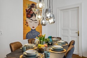 A dining area with a table set for four, featuring a modern chandelier, a large floral artwork on the wall, and a variety of tableware and glassware.