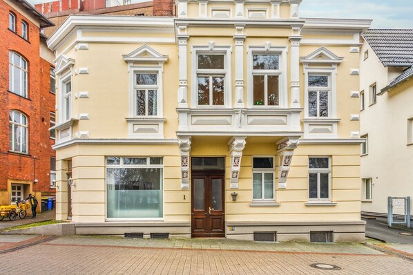 A two-story building with a yellow facade, large windows, and a central entrance with a dark wooden door.