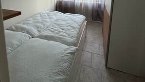 A small, clean room with a bed, a window with sheer curtains, a radiator, and a white refrigerator. - Panorama 180° (Unknown)