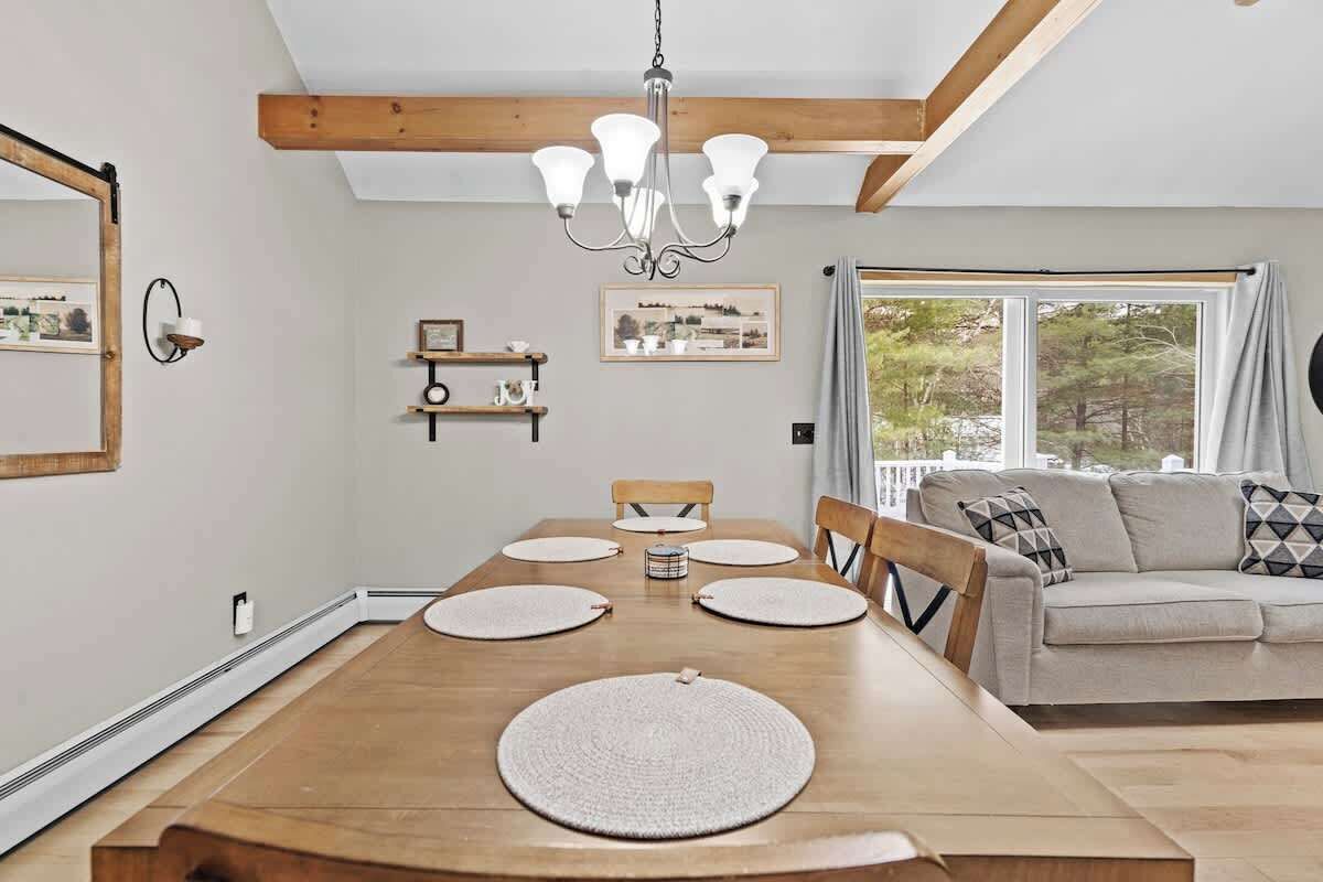 Un comedor con mesa de madera, sillas y una araña. Hay un sofá y una ventana con vista a árboles.