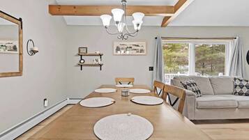Un comedor con mesa de madera, sillas y una araña. Hay un sofá y una ventana con vista a árboles.