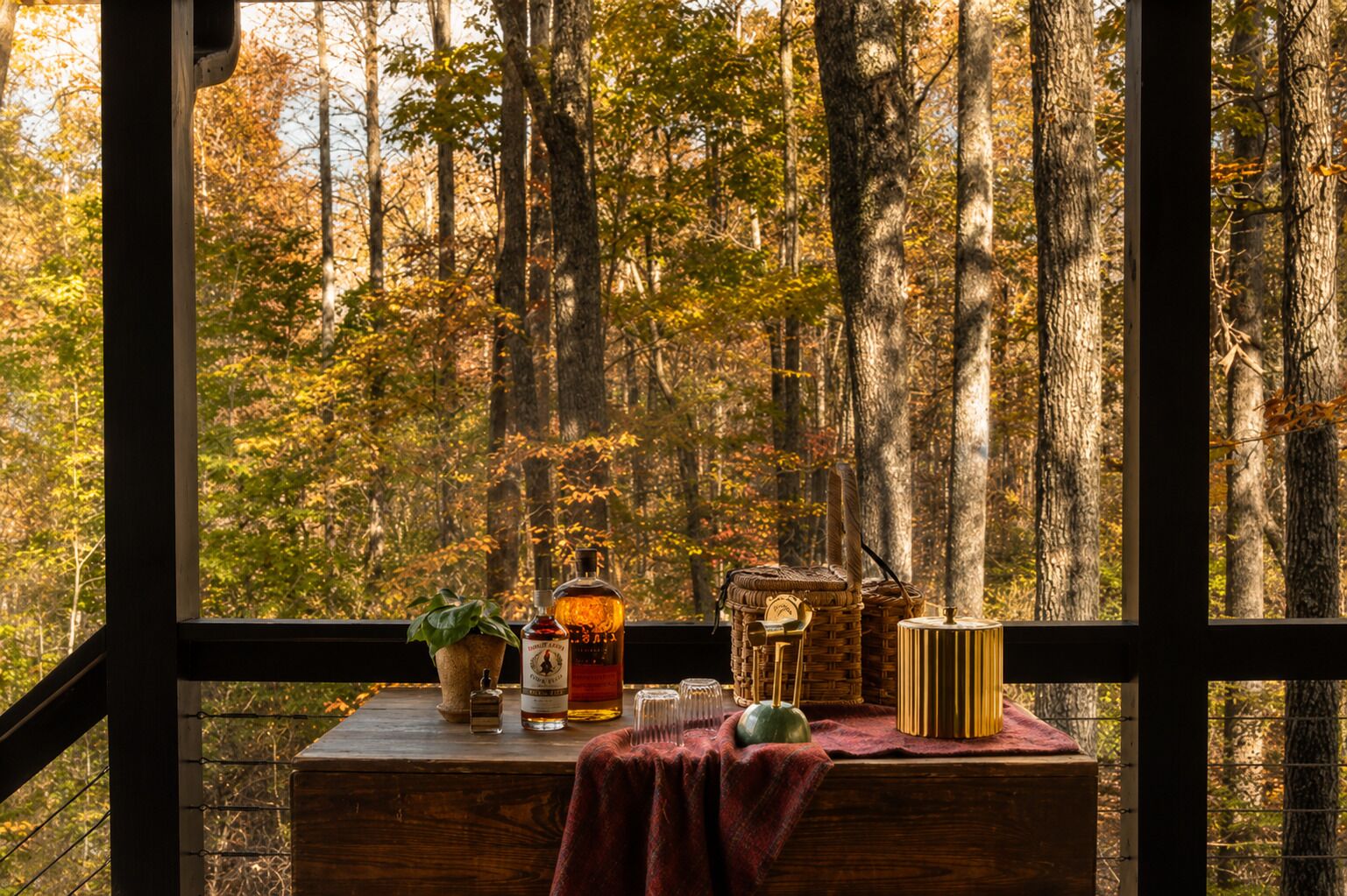 A wooden table with a bottle, glasses, and a plant by a window overlooking a forest.